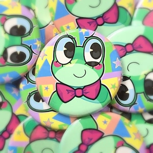 Puede incluir: Un conjunto de botones redondos y coloridos con un diseño de rana de dibujos animados con una pajarita rosa. La rana es de color verde claro con ojos grandes y una expresión sonriente. El fondo de los botones es una mezcla de colores pastel y estrellas.