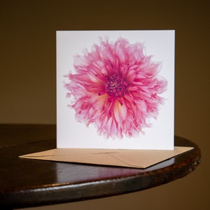 May include: A greeting card featuring a vibrant pink dahlia flower illustration. The card is square and rests on a brown envelope. The dahlia has multiple layers of delicate petals, with a darker center. The card is on a dark wooden surface.