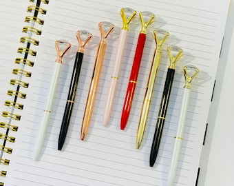 May include: An assortment of diamond-topped pens in various colors, including white, black, rose gold, pink, red, and gold, are arranged on a lined notebook. The pens have a gold or silver barrel and a clear, faceted diamond-shaped top.