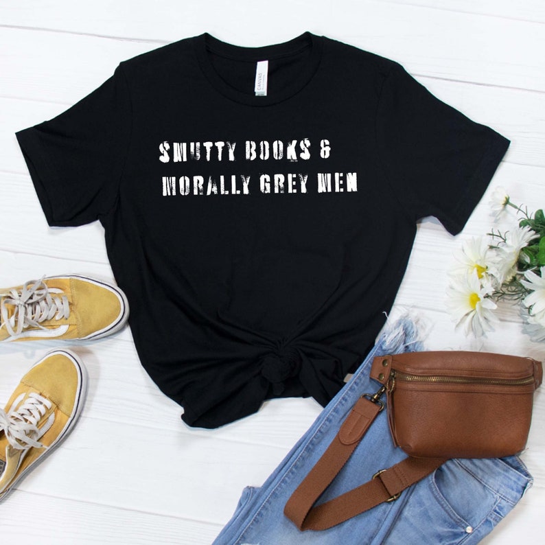 Puede incluir: Camiseta negra con texto blanco que dice "SMUTTY BOOKS & MORALLY GREY MEN". La camiseta est&aacute; colocada sobre una superficie de madera blanca con un par de zapatillas amarillas, unos vaqueros azules y una ri&ntilde;onera marr&oacute;n.