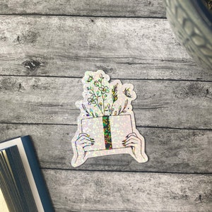 May include: A holographic sticker of a book with flowers growing out of it. The book is being held by two hands. The sticker has a glittery finish.