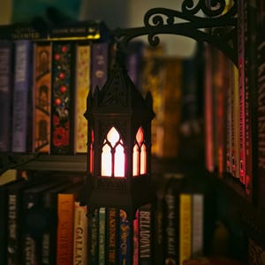 May include: A black, gothic-style hanging lantern with pointed arches and a warm, glowing orange light inside. The lantern is suspended from a decorative, ornate black metal wall bracket, positioned in front of a shelf filled with various books.