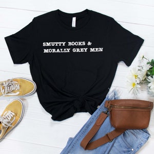 Puede incluir: Camiseta negra con texto blanco que dice "SMUTTY BOOKS & MORALLY GREY MEN". La camiseta est&aacute; colocada sobre una superficie de madera blanca con un par de zapatillas amarillas, unos vaqueros azules y una ri&ntilde;onera de cuero marr&oacute;n.