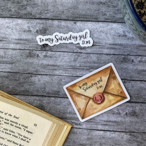 May include: A sticker with a watercolor illustration of a brown envelope with a red wax seal. The text "To my Saturday girl D.M." is written on the envelope.