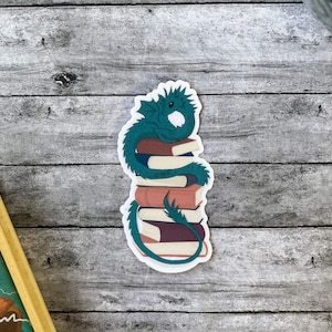 May include: A sticker featuring a teal dragon curled around a stack of books. The books are in shades of brown and white, with the dragon's tail wrapping around them. The sticker has a white border and is set against a wooden background.