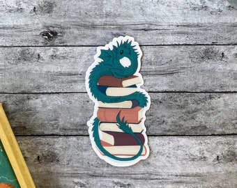 May include: A sticker depicting a teal dragon coiled around a pile of books. The books are various shades of brown and white. The dragon's tail wraps around the books. The sticker has a white border and is on a wooden surface.