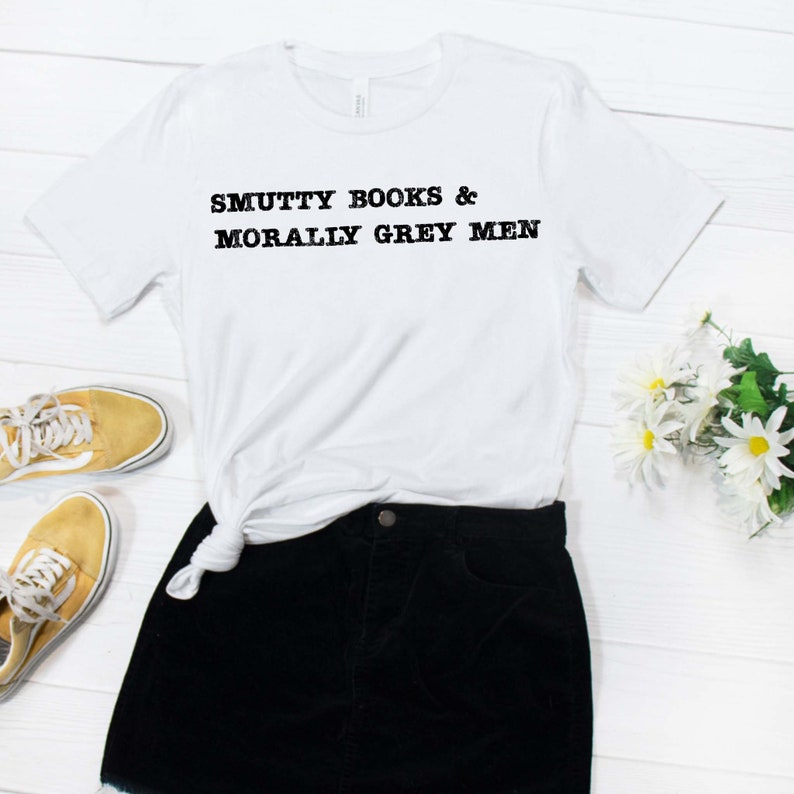 Puede incluir: Camiseta blanca con texto negro que dice "SMUTTY BOOKS & MORALLY GREY MEN". La camiseta est&aacute; atada a la cintura de una falda negra. La falda est&aacute; sobre una superficie de madera blanca.