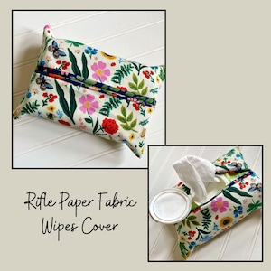 May include: A white fabric tissue box cover with a floral print featuring pink, red, yellow, and blue flowers, green leaves, and butterflies. The text "Rifle Paper Fabric Wipes Cover" is printed below the cover.