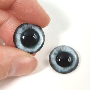May include: Two round, glass doll eyes with a light blue iris and a black pupil. One eye is held by a hand, and the other eye is resting on a white surface. The eyes have a glossy finish.