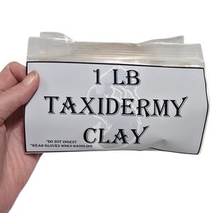 Puede incluir: Un paquete de arcilla para taxidermia, etiquetado como "1 LB TAXIDERMY CLAY" en texto negro en negrita. El paquete se sostiene en una mano y está sellado con plástico transparente. El texto "DO NOT INGEST" y "WEAR GLOVES WHEN HANDLING" también está impreso en el paquete.