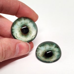 May include: Two green glass eyes with black pupils and white irises. The eyes have a realistic, detailed design.
