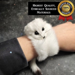 May include: A white fur wrist accessory shaped like a small animal with black eyes, a black nose, and a long tail with a dark brown tip. The accessory features the text Highest Quality, Ethically Sourced Materials and a gold 100% money back seal.