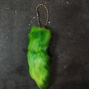 Puede incluir: Un llavero de peluche verde con forma de pata con una cadena dorada.