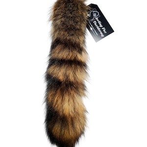 May include: A taxidermy fox tail with a tag. The tail features a mix of brown, black, and tan fur. The tag is black with white text and a logo. The tail is a natural product.