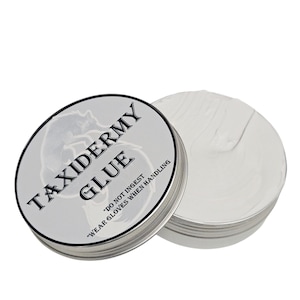 Puede incluir: Un bote redondo de pegamento para taxidermia plateado con tapa blanca. El bote tiene la inscripción "TAXIDERMY GLUE", junto con las advertencias "DO NOT INGEST" y "WEAR GLOVES WHEN HANDLING". El pegamento del interior es blanco.