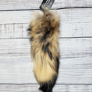 May include: A natural fox tail, featuring a mix of brown, black, and cream-colored fur. The tail is attached to a small tag that reads "Sterling Fox Taxidermy". The tail is displayed against a white wooden background.
