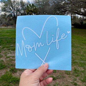 Puede incluir: Una calcoman&iacute;a de vinilo cuadrada de color azul claro que presenta el texto Mom life en cursiva blanca con una forma de coraz&oacute;n integrada en las letras y un peque&ntilde;o icono de coraz&oacute;n al final de la palabra life, seguido de una x estilizada.