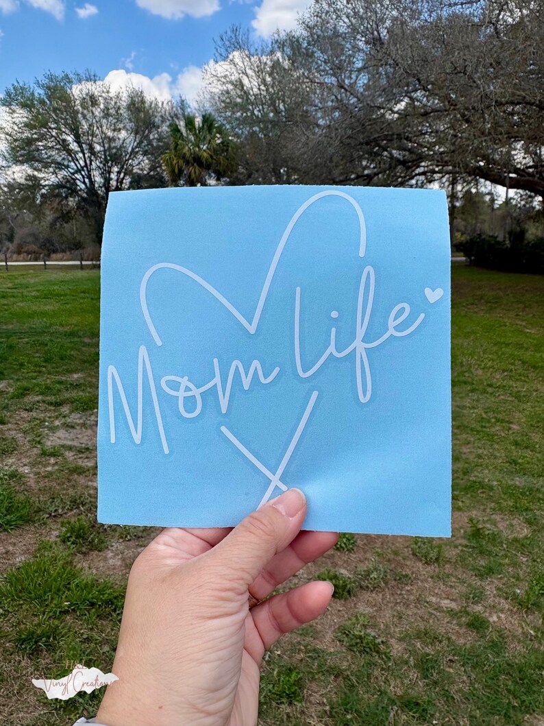 Puede incluir: Una calcoman&iacute;a de vinilo cuadrada de color azul claro que presenta el texto Mom life en cursiva blanca con una forma de coraz&oacute;n integrada en las letras y un peque&ntilde;o icono de coraz&oacute;n al final de la palabra life, seguido de una x estilizada.