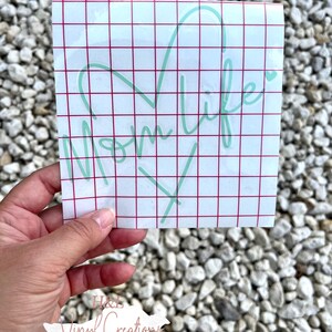Puede incluir: Una calcoman&iacute;a de vinilo cuadrada con texto en letra cursiva de color verde menta claro que dice Mom life junto a un contorno en forma de coraz&oacute;n. El dise&ntilde;o est&aacute; sobre un papel protector blanco con una cuadr&iacute;cula roja para alineaci&oacute;n.