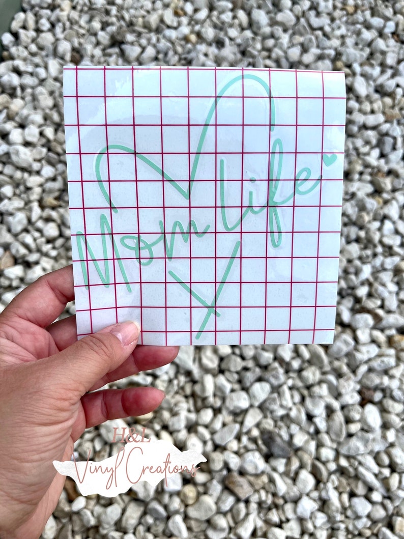 Puede incluir: Una calcoman&iacute;a de vinilo cuadrada con texto en letra cursiva de color verde menta claro que dice Mom life junto a un contorno en forma de coraz&oacute;n. El dise&ntilde;o est&aacute; sobre un papel protector blanco con una cuadr&iacute;cula roja para alineaci&oacute;n.