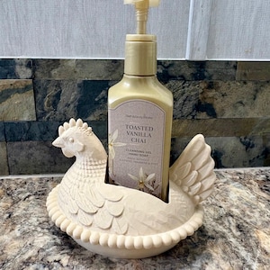 May include: A cream-colored chicken-shaped soap dispenser holder with a bottle of hand soap. The hand soap bottle is labeled "TOASTED VANILLA CHAI" and "CLEANSING GEL HAND SOAP". The holder is made of ceramic or a similar material.
