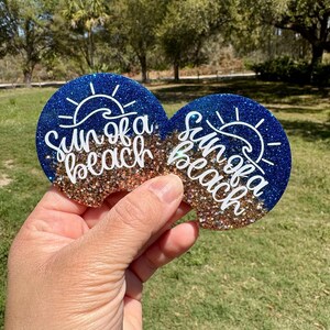 May include: Two round car coasters with a blue glitter top and a tan glitter bottom. The coasters have the words "Sun of a beach" in white script with a sun graphic. The background is a grassy area with trees.