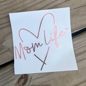 Puede incluir: Una calcoman&iacute;a cuadrada blanca con el texto Mom life en una fuente cursiva de color oro rosa met&aacute;lico. El dise&ntilde;o incluye una forma de coraz&oacute;n integrada con las letras y un peque&ntilde;o icono de coraz&oacute;n sobre la letra i. La calcoman&iacute;a tiene un acabado liso.