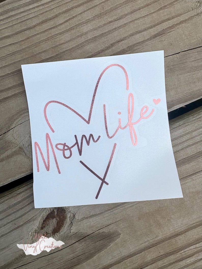 Puede incluir: Una calcoman&iacute;a cuadrada blanca con el texto Mom life en una fuente cursiva de color oro rosa met&aacute;lico. El dise&ntilde;o incluye una forma de coraz&oacute;n integrada con las letras y un peque&ntilde;o icono de coraz&oacute;n sobre la letra i. La calcoman&iacute;a tiene un acabado liso.