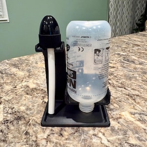 May include: A black plastic stand holding a clear plastic bottle and a black nozzle. The bottle has white text and a label. The stand is on a countertop with a marble pattern. The nozzle has a white tube.