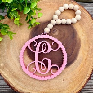 May include: A pink circular ornament featuring a decorative script letter C in the center, surrounded by a scalloped border. The ornament is attached to a loop of natural light-colored wooden beads. The text Vinyl Creations is visible.