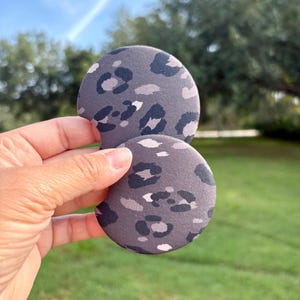 May include: Two gray car coasters with a leopard print design. The coasters are round and made of a soft material. The background is a blurred view of green grass and trees.