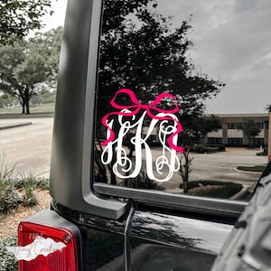 May include: A white vinyl monogram decal featuring the letters H and K in a decorative script font, topped with a bright pink bow graphic. The decal is applied to the rear window of a black vehicle, positioned above the red taillight.