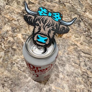 May include: A wooden Highland cow can topper with a turquoise bow and accents, placed on top of a Dr. Pepper can. The cow has a brown body, black outline, and white horns. The can is silver with red text.