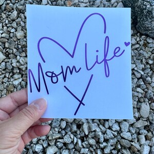 Puede incluir: Una calcoman&iacute;a cuadrada blanca con el texto morado Mom life en una fuente cursiva. El dise&ntilde;o incluye un gran contorno de coraz&oacute;n morado sobre el texto y un peque&ntilde;o icono de coraz&oacute;n morado al final de la palabra life, con una X morada debajo.