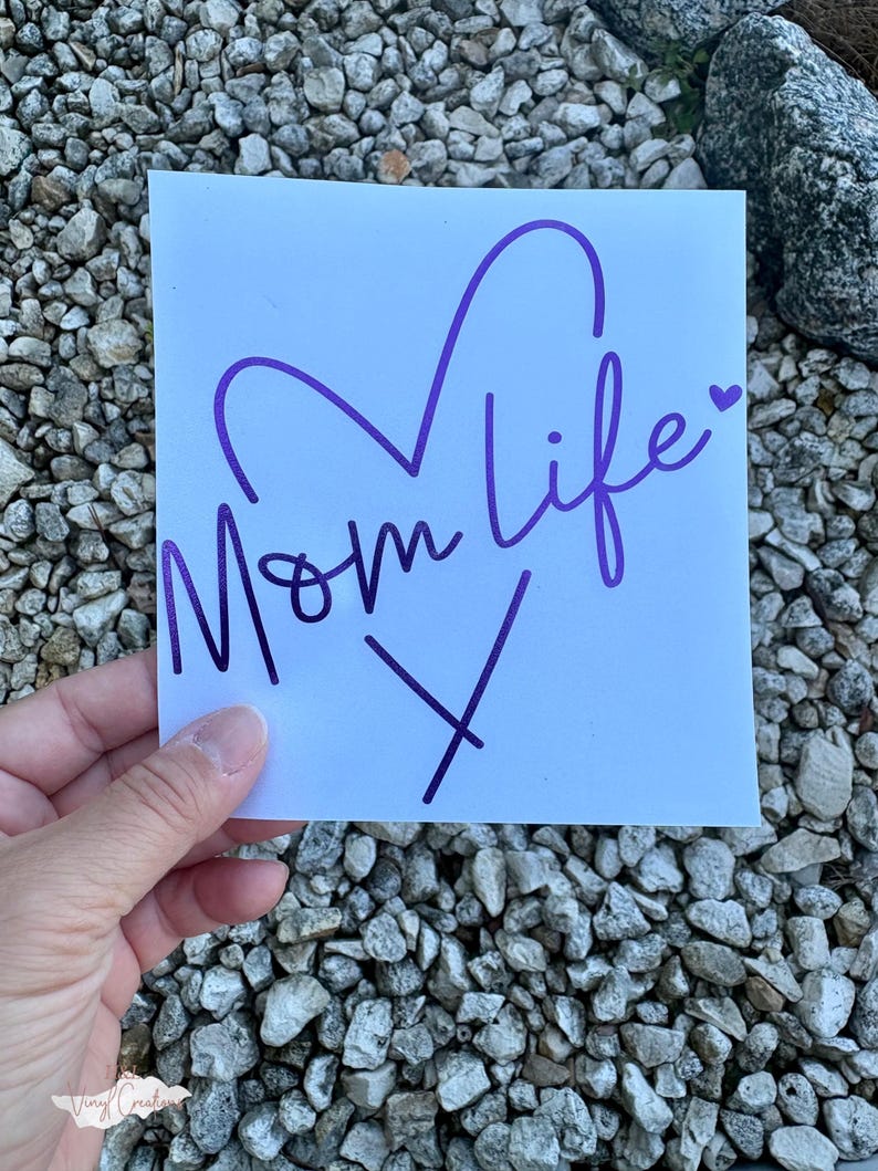 Puede incluir: Una calcoman&iacute;a cuadrada blanca con el texto morado Mom life en una fuente cursiva. El dise&ntilde;o incluye un gran contorno de coraz&oacute;n morado sobre el texto y un peque&ntilde;o icono de coraz&oacute;n morado al final de la palabra life, con una X morada debajo.