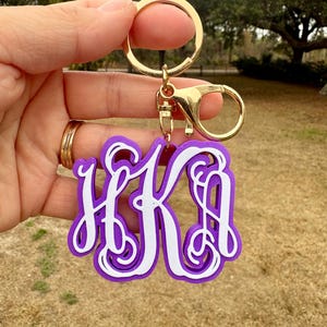 May include: A purple and white monogram keychain featuring the letters HKA in a script font. The keychain is attached to a gold-toned metal split ring and a lobster claw clasp. The design consists of a white script monogram layered over a purple base.