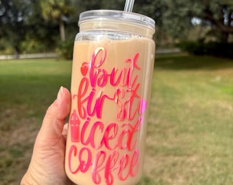 May include: A clear mason jar style tumbler filled with iced coffee, featuring a pink and iridescent vinyl decal that reads "but first iced coffee." The tumbler has a clear lid and a clear straw. The background is a blurred outdoor scene.