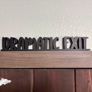 May include: Black wooden sign that reads "DRAMATIC EXIT" in a decorative font. The sign is placed on a wooden surface against a light-colored wall. The sign is a decorative item.
