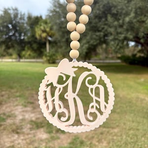 May include: A light beige circular ornament featuring a cutout monogram with the letters HKA, topped with a small bow. The ornament is suspended from a string of smooth, round, light-colored wooden beads, creating a decorative hanging piece.