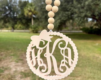 May include: A light beige circular ornament featuring a cutout monogram with the letters HKA, topped with a small bow. The ornament is suspended from a string of smooth, round, light-colored wooden beads, creating a decorative hanging piece.