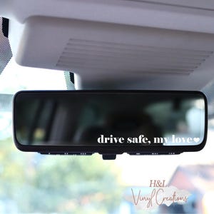 May include: A rectangular rearview mirror featuring a white vinyl decal with the text "drive safe, my love" followed by a small heart symbol. The lettering is applied directly to the surface of the mirror, creating a clear and legible message.