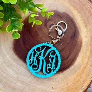May include: A circular turquoise keychain featuring an intricate, stylized monogram of the letters H, K, and A. The monogram is enclosed within a scalloped border and attached to a silver-toned metal split ring and a lobster claw clasp.