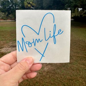 Puede incluir: Una calcoman&iacute;a de vinilo blanca cuadrada con el texto azul Mom life escrito en una fuente cursiva. El dise&ntilde;o incluye un contorno de coraz&oacute;n azul colocado sobre el texto y una letra X azul situada debajo del texto sobre el fondo blanco.