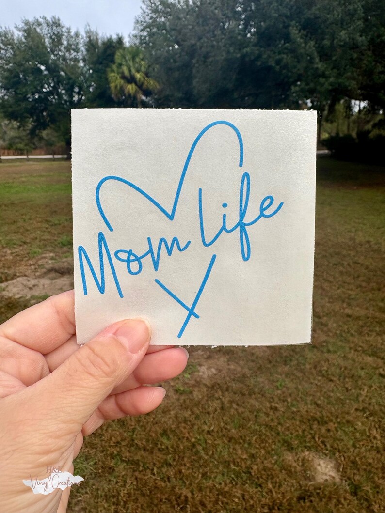 Puede incluir: Una calcoman&iacute;a de vinilo blanca cuadrada con el texto azul Mom life escrito en una fuente cursiva. El dise&ntilde;o incluye un contorno de coraz&oacute;n azul colocado sobre el texto y una letra X azul situada debajo del texto sobre el fondo blanco.