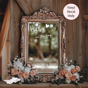 May include: A gold ornate framed mirror with the words "Baby on the way" and "October 2025" in white script. The mirror is surrounded by bouquets of peach and white flowers. A pink circle with the text "Vinyl Decal Only" is in the upper right corner.