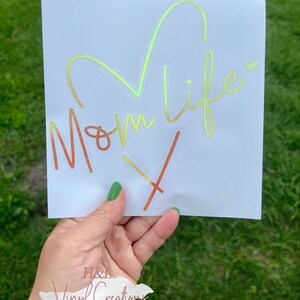 Puede incluir: Una calcoman&iacute;a cuadrada blanca con el texto Mom Life en una fuente cursiva con un degradado de color que va del naranja al amarillo y al verde. Un gran contorno de coraz&oacute;n est&aacute; integrado sobre el texto y un peque&ntilde;o coraz&oacute;n a la derecha.