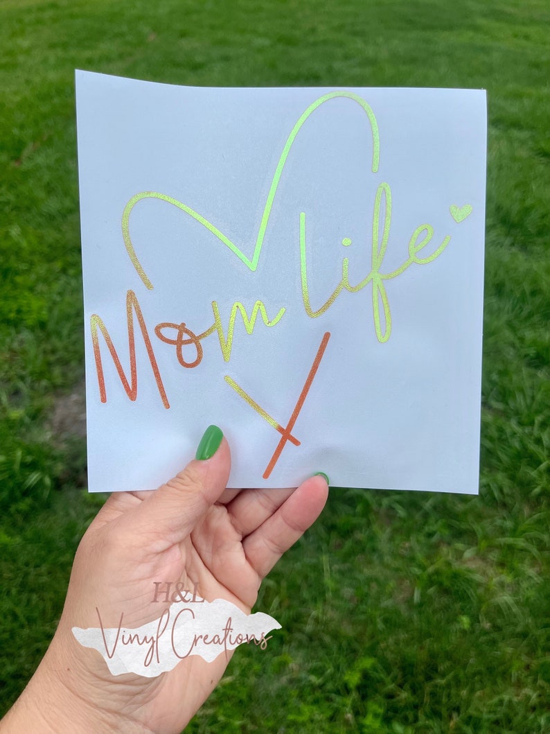 Puede incluir: Una calcoman&iacute;a cuadrada blanca con el texto Mom Life en una fuente cursiva con un degradado de color que va del naranja al amarillo y al verde. Un gran contorno de coraz&oacute;n est&aacute; integrado sobre el texto y un peque&ntilde;o coraz&oacute;n a la derecha.