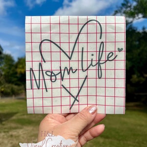 Puede incluir: Una calcoman&iacute;a de vinilo cuadrada que presenta el texto cursivo negro Mom life con una forma de coraz&oacute;n integrada en las letras. La calcoman&iacute;a est&aacute; aplicada sobre una hoja de soporte blanca impresa con un patr&oacute;n de cuadr&iacute;cula roja.