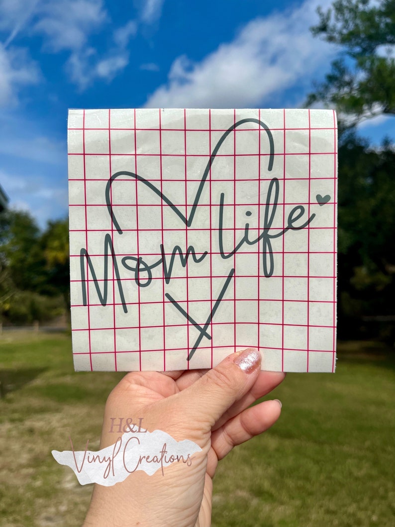 Puede incluir: Una calcoman&iacute;a de vinilo cuadrada que presenta el texto cursivo negro Mom life con una forma de coraz&oacute;n integrada en las letras. La calcoman&iacute;a est&aacute; aplicada sobre una hoja de soporte blanca impresa con un patr&oacute;n de cuadr&iacute;cula roja.