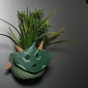 Puede incluir: Una maceta de cerámica verde con forma de cara de monstruo de dibujos animados con una pequeña planta que crece en la parte superior.